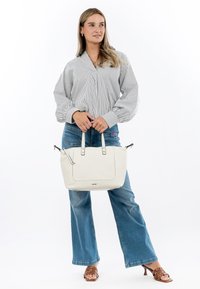 Sac à main en cuir blanc avec deux anses supérieures, fermeture éclair et poche latérale. Le modèle porte un chemisier rayé et un jean large bleu.