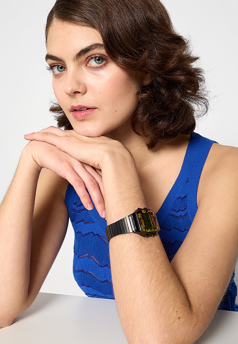 Reloj de metal negro con una carátula rectangular, pantalla digital colorida, colocado sobre un modelo que lleva un top de punto texturizado azul.