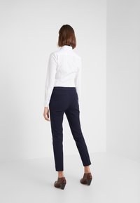 Biała koszula z długim rękawem, granatowe dopasowane spodnie, brązowe buty na obcasie. Gładka tkanina, krój na miarę, z tylnymi kieszeniami w spodniach.