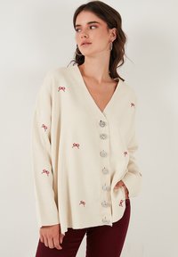 Cardigan oversize color crema con scollo a V, decorato con ricami a forma di fiocco rosso e grandi bottoni argentati sul davanti. Tessuto morbido e testurizzato.