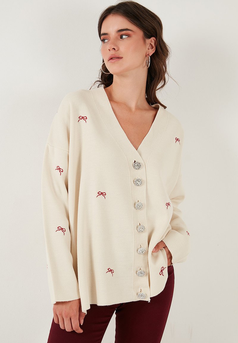 Cardigan oversize color crema con scollo a V, decorato con ricami a forma di fiocco rosso e grandi bottoni argentati sul davanti. Tessuto morbido e testurizzato.