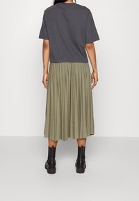 Grijze top met korte mouwen gecombineerd met een geplooide olijfgroene midi-rok en zwarte enkellaarsjes, gezien van achteren.