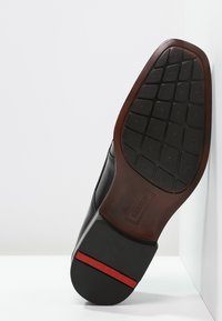 Chaussure en cuir noir avec une semelle en caoutchouc texturée présentant des motifs rectangulaires et une bande d'accent rouge sur le bord inférieur.