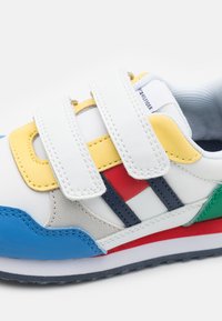 Baskets blanches avec des sangles Velcro, ornées d'accents jaunes, bleus, rouges et verts, et du logo Tommy Hilfiger sur la languette.