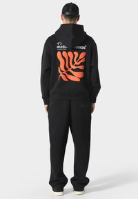 Černá mikina s velkým oranžovým grafickým motivem listu na zadní straně a textem "IRASUT Studios". V kombinaci se shodnými černými tepláky. Uvolněný střih.