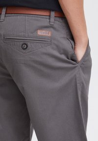 Graue Hose mit einer glatten Textur und einer Gesäßtasche mit einem schwarzen Knopf. Abgerundet mit einem braunen Lederpatch, das "Isolid" trägt.