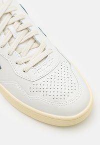 Weiße Ledersneaker mit perforierter Zehenkappe, hellbeiger Sohle und cremfarbenen Schnürsenkeln. Minimalistisches Design mit kontrastierenden seitlichen Details.
