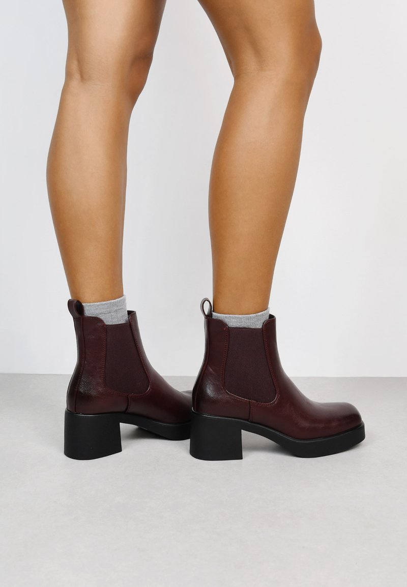 Bottines en cuir bordeaux à talons blocs, avec des panneaux élastiques sur les côtés et des tirettes, portées avec des chaussettes grises, affichant une texture lisse.
