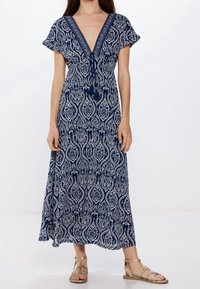 Robe maxi bleu marine avec motif cachemire blanc, encolure en V, manches courtes et accent à pampilles à la taille. Longueur jusqu'aux chevilles.