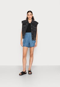 Svart denims ärmlös jacka över en svart topp, i kombination med ljusblå högmidjade shorts och svarta sandaler. Enkel, minimalistisk look.