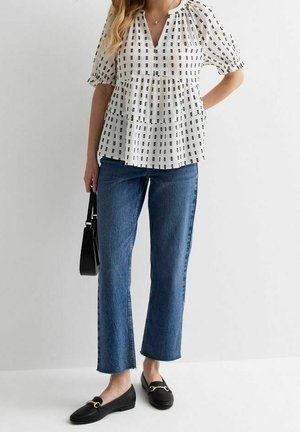 Mujer que lleva una blusa blanca con estampado y mangas abullonadas, vaqueros azules recortados, mocasines negros y lleva un bolso negro.