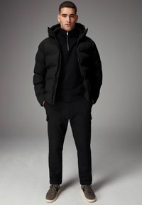 Sort pufferjakke med høj krave og lynlås, lagd ovenpå en sort sweatshirt. Sammen med sorte joggers og grå sneakers.