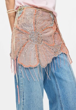 Stor gennemsigtig lyserød blomsterapplikation med palietter og perlebroderi på lyseblå denimjeans, bundet med beige snore i taljen.