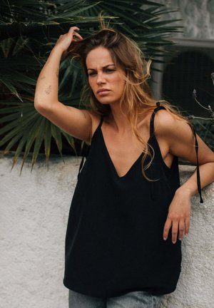Woman with long hair in black sleeveless top leaning on white wall, raising one arm to touch her hair, with green palm leaves in background.