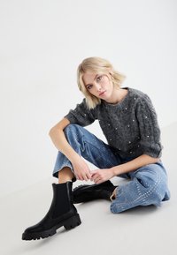 Bottines noires avec des semelles en caoutchouc texturé, associées à un pull en maille gris orné de perles et à un jean en denim clair, brodé.
