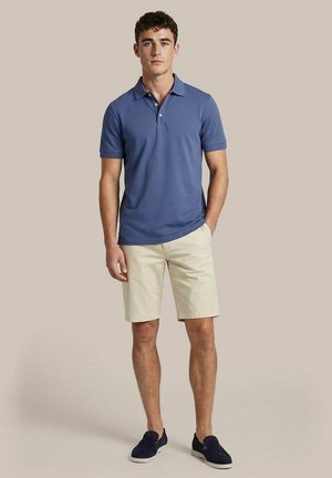 Jonge man staat met handen in de zakken, draagt een blauw poloshirt, beige korte broek en donkerblauwe slip-on schoenen op een effen achtergrond.