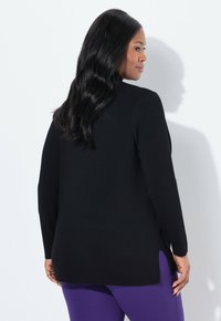 Vrouw met lang zwart haar, gekleed in een zwart shirt met lange mouwen en paarse broek, die zijwaarts staat tegen een witte muur met haar schaduw zichtbaar.