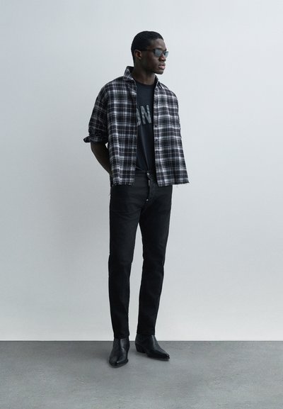 Άνδρας με μαύρα γυαλιά ηλίου, μαύρο τζιν, μαύρες μπότες, μαύρο γραφικό μπλουζάκι και ανοιχτό πουκάμισο με μοτίβο ασπρόμαυρο καρό, στέκεται με τα χέρια πίσω από την πλάτη.