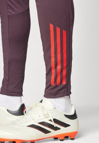 Pantalones deportivos morados con tres rayas rojas verticales en la pierna izquierda, combinados con zapatillas blancas que tienen acentos negros y suelas naranjas.