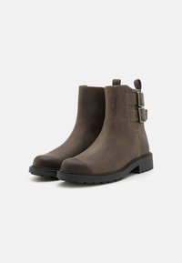 Botas marrones de tobillo con un acabado de cuero suave, punta redonda, suela de goma negra gruesa y dos hebillas decorativas a los lados.