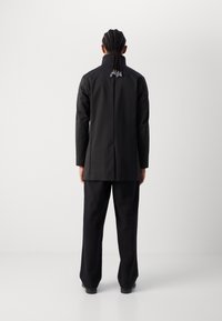Zwarte stoffen blazer met hoge kraag, slim fit en een ventilerende achterkant. Gecombineerd met losse zwarte broek. Achteraanzicht met zichtbare gevlochten haarsstijl.