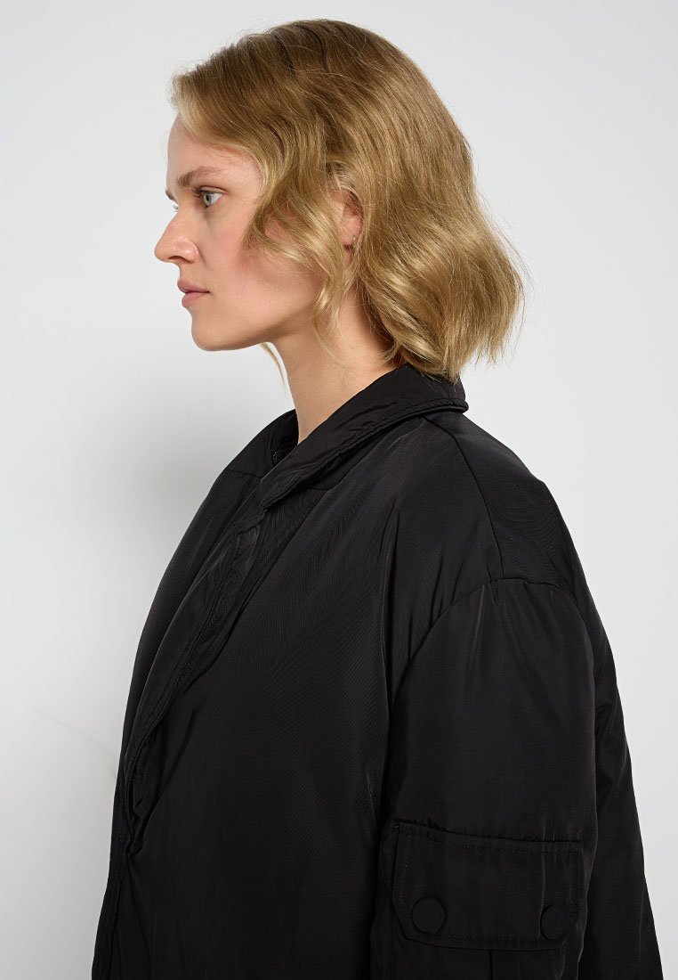 Schwarze gepolsterte Jacke mit hohem Kragen, strukturierter Oberfläche und einer knöpfbaren Tasche am Ärmel. Weicher, leichter Stoff; oversized Passform.