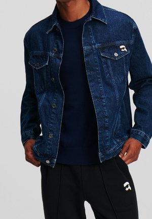 Homme portant une veste en jean bleu foncé sur un pull marine et un pantalon noir avec un petit logo blanc de visage sur la poche de la veste et le pantalon.