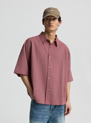 Giovane uomo con berretto beige "Las Vegas", camicia rossa a quadri con maniche corte e bottoni e jeans blu su uno sfondo chiaro e uniforme.