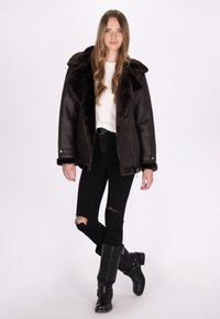 Giacca in shearling marrone con colletto ampio, jeans neri strappati e stivali neri fino al ginocchio. La giacca presenta accenti metallici e una texture morbida.