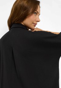 Frau mit hellbraunem Haar, die ein lockeres schwarzes Oberteil trägt und seitlich schaut, mit ihrem Arm nahe ihrer Schulter erhoben, vor einem weißen Hintergrund.