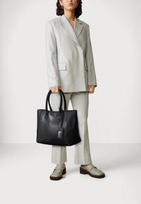 Czarna skórzana torba typu tote z teksturą wytłaczanego krokodyla, podwójne uchwyty na górze oraz zawieszka; zestawiona z jasnoszarym garniturem i jasnymi butami.