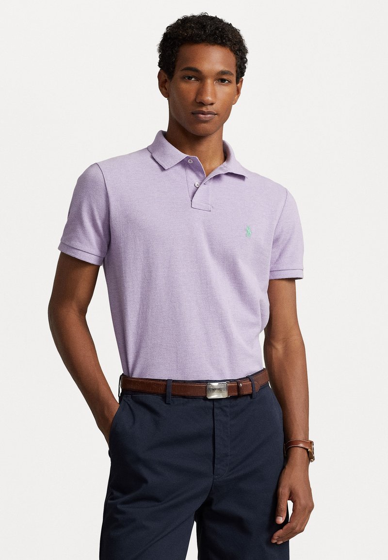 Lavendli lühikese varrukaga polo särk nööpidega krae ja rohelise embroideritud logoga, kombineeritud tumedate pükstega ja pruunist nahkrihmaga.