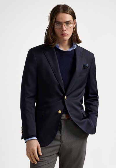 Blazer blu marina con bottoni dorati, abbinato a un maglione blu marina e camicia azzurro chiaro; pantaloni grigi; foulard con pois bianchi.