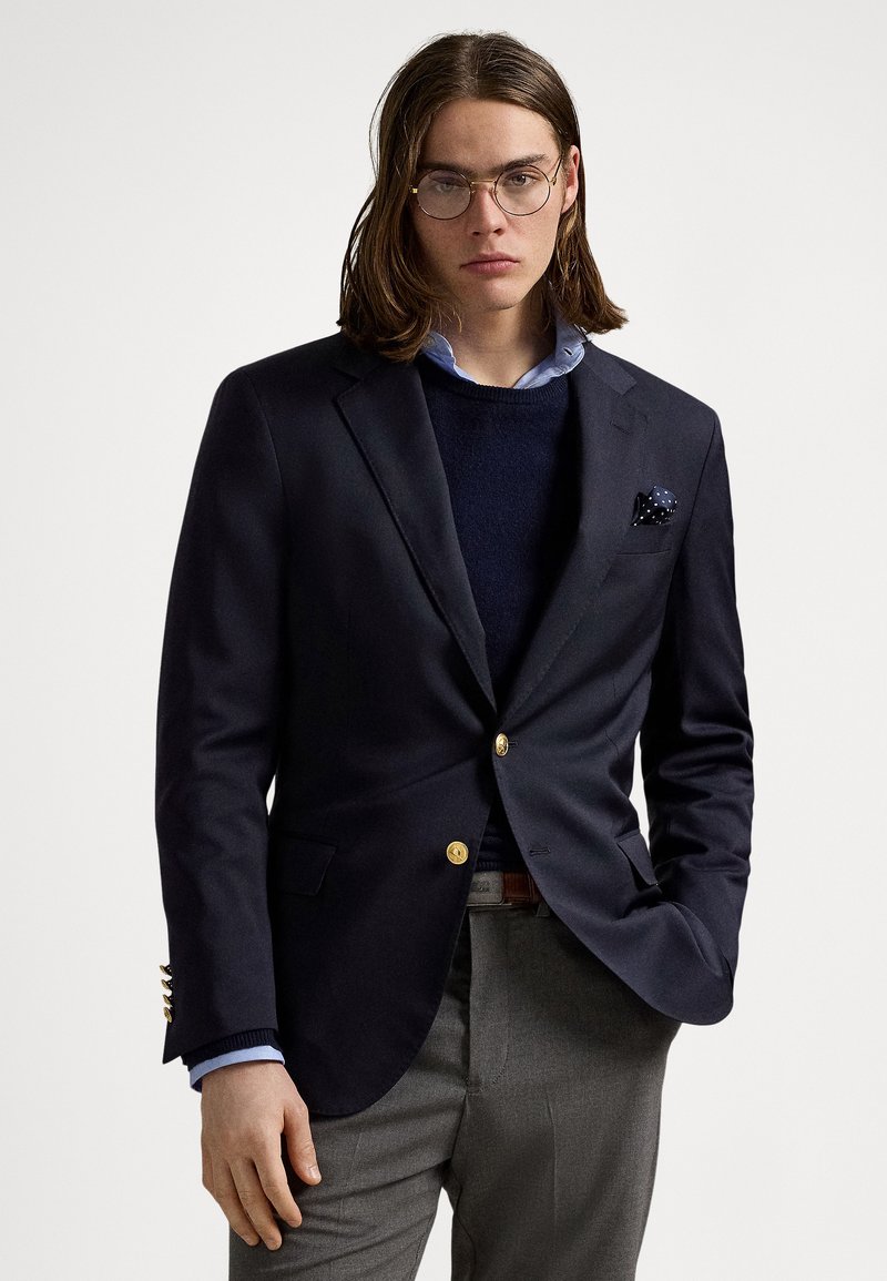 Blazer azul-marinho com botões dourados, combinado com um suéter azul-marinho e camisa azul-claro; calças cinzentas; lenço de bolso com bolinhas brancas.