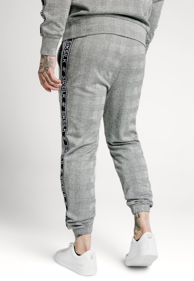 Harmaat ruudulliset jogginghousut mustilla sivunauhoilla, joissa on "SIKSILK"-logo. Joustavat nilkat ja pehmeä rakenne, yhdistettynä valkoisiin lenkkareihin.