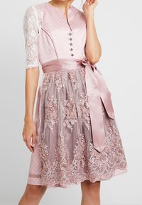 Robe rose avec un corsage texturé, des manches en dentelle et une jupe brodée. Présente une ceinture en satin et des détails de boutons à l'avant.