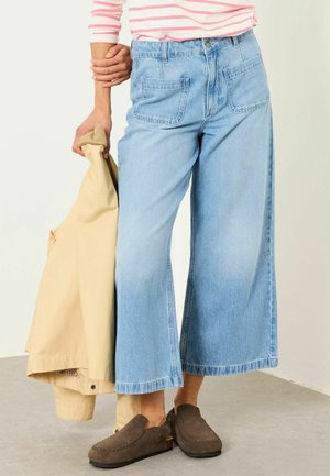 Osoba w krótkich jasnoniebieskich dżinsach o szerokich nogawkach, brązowych wsuwanych butach i paskowanej różowo-białej bluzce, trzymająca beżowy płaszcz w lewej ręce.