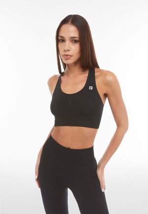 Schwarzer Sport-BH mit breiten Trägern, glatter Oberfläche und einem Logopatch. Kombiniert mit hoch taillierten schwarzen Leggings. Minimalistisches und enganliegendes Design.
