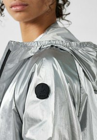 Zilveren windjack met een glanzende afwerking, elastische capuchon en een zwart logovlekje op de linker schouder. Soepele textuur, lichtgewicht ontwerp.