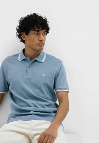 Polo di colore azzurro chiaro con pattina a tre bottoni, bordature bianche sul colletto e sulle maniche, caratterizzato da un piccolo logo ricamato sul petto.