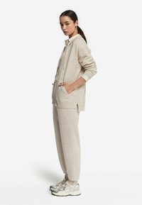 Beige huva sweatshirt med fickor och sidokilar, parat med matchande joggare. Modellen bär sneakers med silverdetaljer.