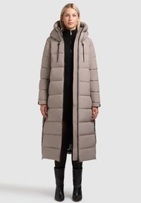 Lang, pufferjakke i lyse beige, med hætte, quiltet design, sorte lynlåsdetaljer, og en lige silhuet. Båret over en sort sweaterkjole.