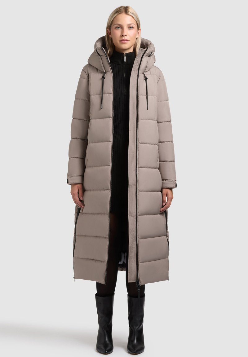Lang, pufferjakke i lyse beige, med hætte, quiltet design, sorte lynlåsdetaljer, og en lige silhuet. Båret over en sort sweaterkjole.
