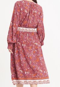 Mekko, jossa on pitkät hihat, vadelmanvärinen sävy, paisley-kuvioita, kokoontuva vyötärö ja valkoinen brodeerattu vyö. Midimittainen, jossa on liehuva hame.