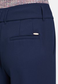 Marineblaue maßgeschneiderte Hose mit strukturiertem Stoff, einer Gesäßtasche und einem kleinen Metall-Logo-Akzent am Bund.