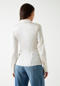 Blusa bianca in satin con una vestibilità strutturata e sartoriale, maniche lunghe e colletto classico, abbinata a jeans in denim azzurri.