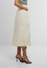 Gonna in denim beige chiaro con tasche, lunghezza alla caviglia, caratterizzata da un taglio dritto e cuciture in vista. Abbinata a sandali neri con tacco alto e cinturini.
