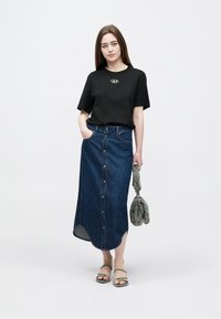 Juoda trumpomis rankovėmis marškinėliai su logotipu; mėlynos spalvos džinsinė sijonas su sagomis; pilka rankinė; minimalistinės sandalės. Sijonas turi aukštą skeltuką ir kišenes.