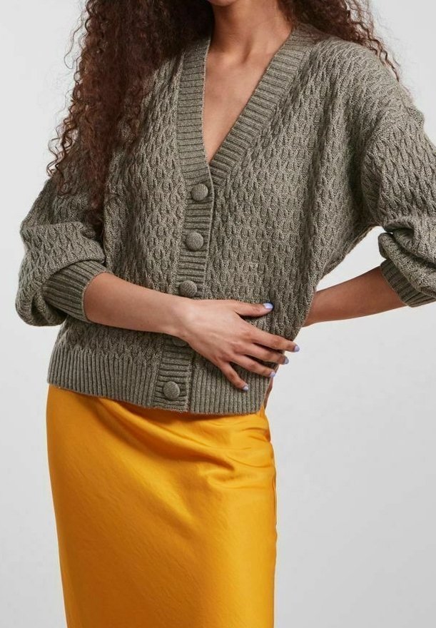 Nainen, jolla on päällään oliivinvihreä palmikkoneuleinen napillinen neuletakki, jonka hihat on kääritty, ja kirkkaan keltainen satiinishortsi, käsi puolellaan.