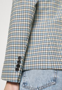 Maniche di blazer in plaid beige, blu e nero con quattro bottoni neri. Il tessuto ha una trama testurizzata, che mostra un classico motivo a quadri.